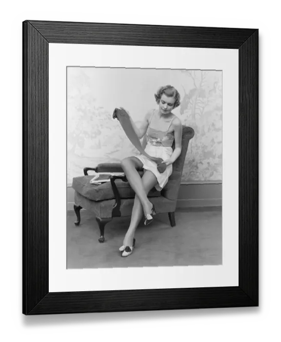 Framed Print : Woman wearing lingerie, sitting on arm of chair, holding up pair of silk stockings