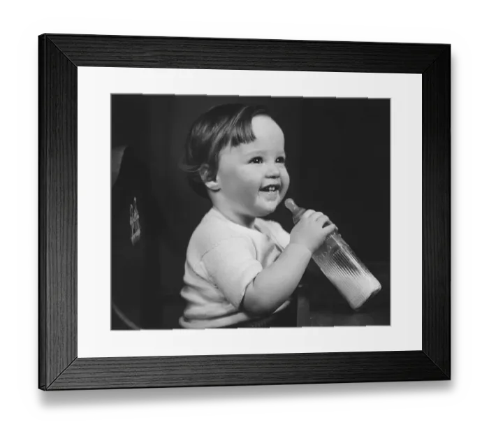 Framed Print : Baby girl (9-12 months) sitting in highchair, holding bottle of milk (B&W) Framed Print : Baby girl (9-12 months) sitting in highchair, holding bottle of milk (B&W)