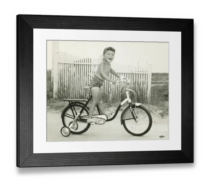 Framed Print : Girl (6-7) riding bicycle with side wheels, (B&W) Framed Print : Girl (6-7) riding bicycle with side wheels, (B&W)