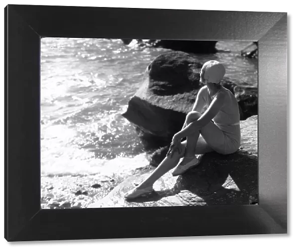 Lorelei. circa 1930: A swimsuited woman sits on a rock gazing out to sea