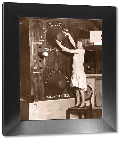 Young woman, standing on chair, adjusting giant radio tuner (B&W)