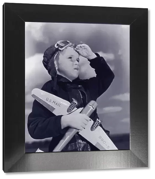 Boy (8-10) wearing flying cap and goggles holding toy plane (B&W)