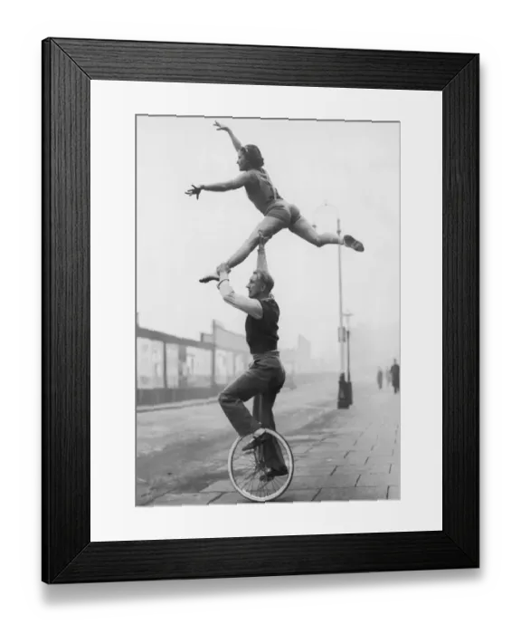 Framed Print : Male acrobat on unicycle supporting woman in air (B&W) Framed Print : Male acrobat on unicycle supporting woman in air (B&W)