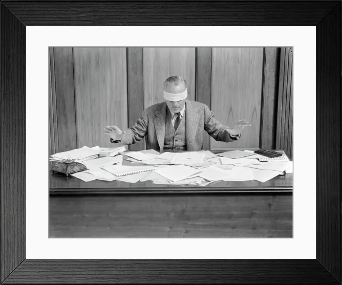 Framed Print : Blindfolded Businessman At Desk Covered With Papers Framed Print : Blindfolded Businessman At Desk Covered With Papers