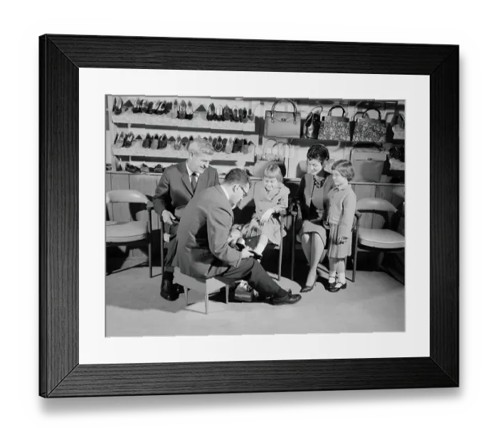 Framed Print : Family of four in shoe shop, salesman fitting shoe on girl as mother, father and sister look Framed Print : Family of four in shoe shop, salesman fitting shoe on girl as mother, father and sister look