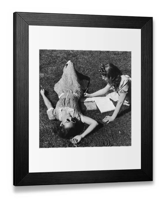 Framed Print : Two teenage girls (14-16) on grass, one doing homework (B&W) Framed Print : Two teenage girls (14-16) on grass, one doing homework (B&W)