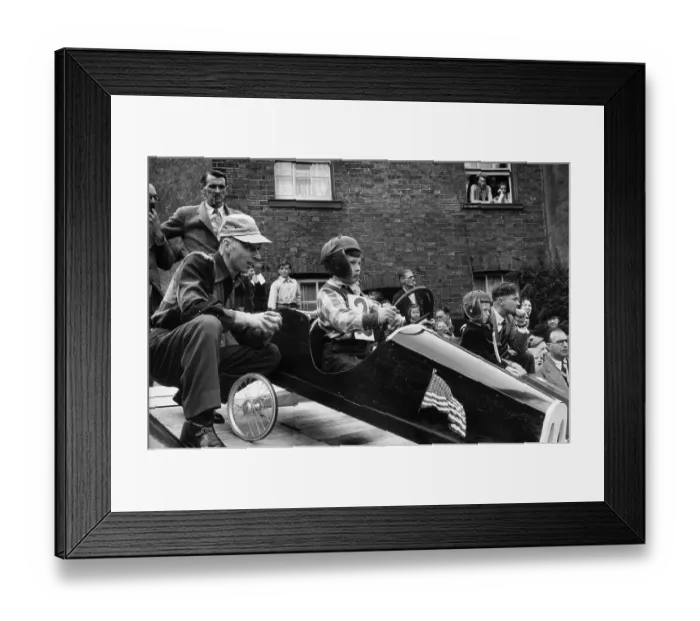 Framed Print : Mini Prix. 12th July 1952: A toddler in a vehicle bearing a US flag waits Framed Print : Mini Prix. 12th July 1952: A toddler in a vehicle bearing a US flag waits