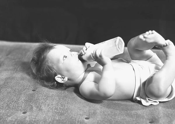 Baby (6-9 months) lying on bed, drinking from bottle, (B&W)