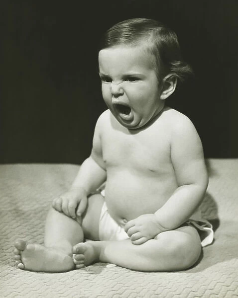 Baby boy (12-18 months) sitting on bedspread, yawning, (B&W)