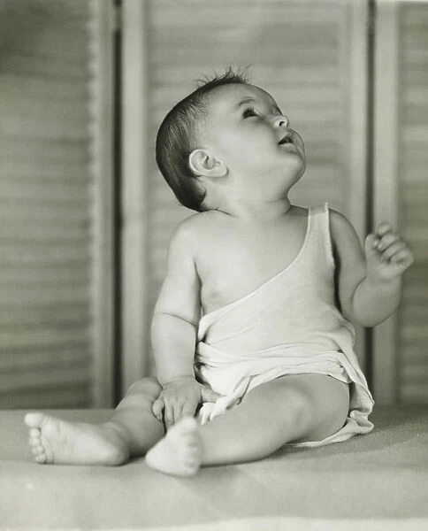 Baby boy (6-12) sitting on floor indoors, looking upwards, (B&W)
