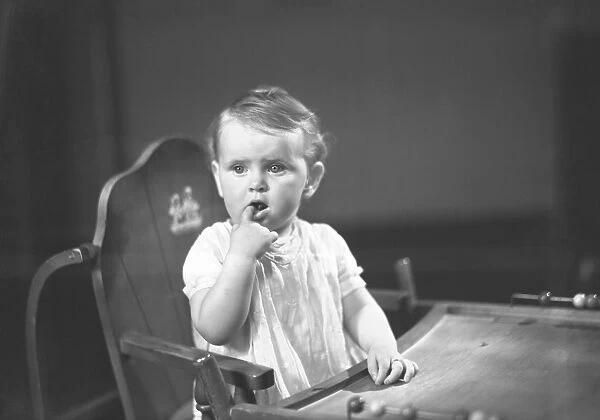 Baby girl (9-12 months) sitting in high chair, holding finger in mouth, (B&W)