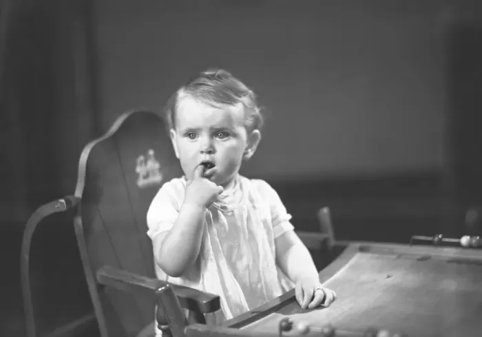 Framed Print : Baby girl (9-12 months) sitting in high chair, holding finger in mouth, (B&W)
