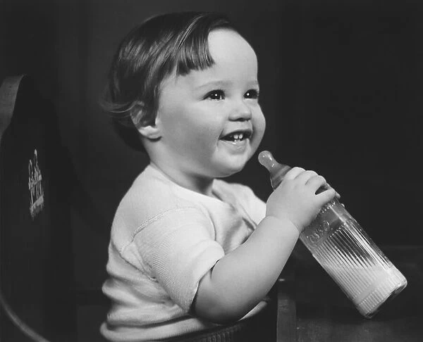 Baby girl (9-12 months) sitting in highchair, holding bottle of milk (B&W)