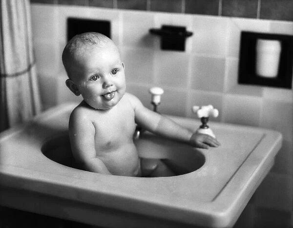 Baby sitting in porcelain sink in bathroom, sticking out tongue. (Photo by H