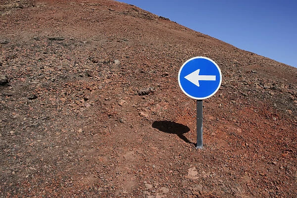 Blue sign with a white arrow pointing left, on red lava, Lanzarote, Canary Islands, Spain