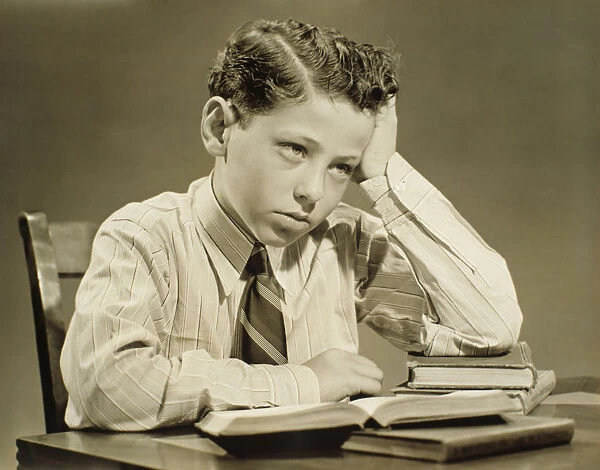 Boy (10-11) sitting at table over open book, head resting on hand, (B&W), close-up