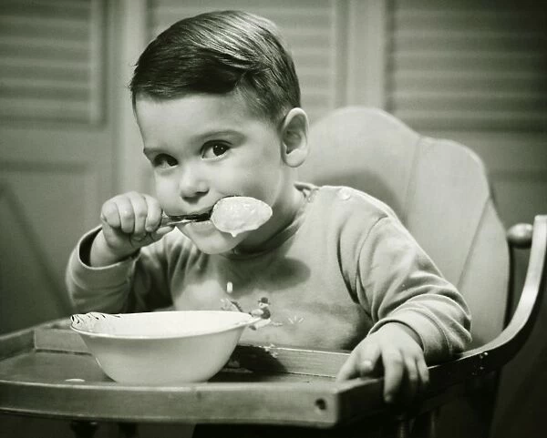 Boy (4-5) sitting in high chair, eating porridge, (B&W), portrait