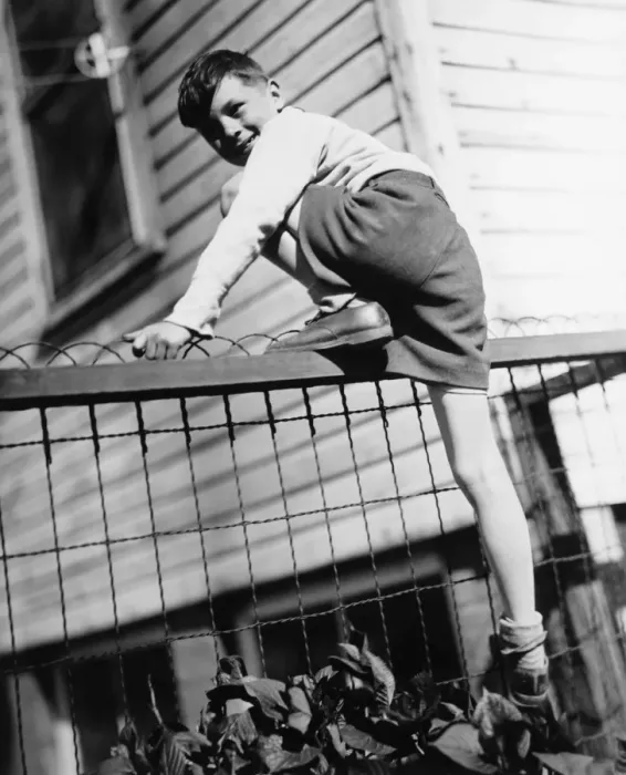 Framed Print : Boy (8-9) climbing on enclosure in front of house, (B&W) Framed Print : Boy (8-9) climbing on enclosure in front of house, (B&W)