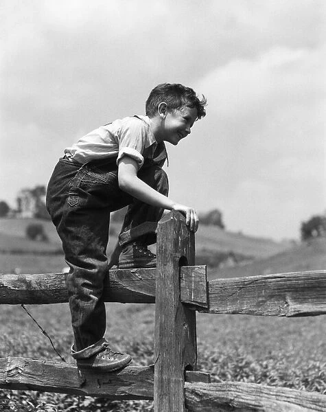Boy in denim bib overalls, climbing over wooden split rail fence on farm. (Photo by H