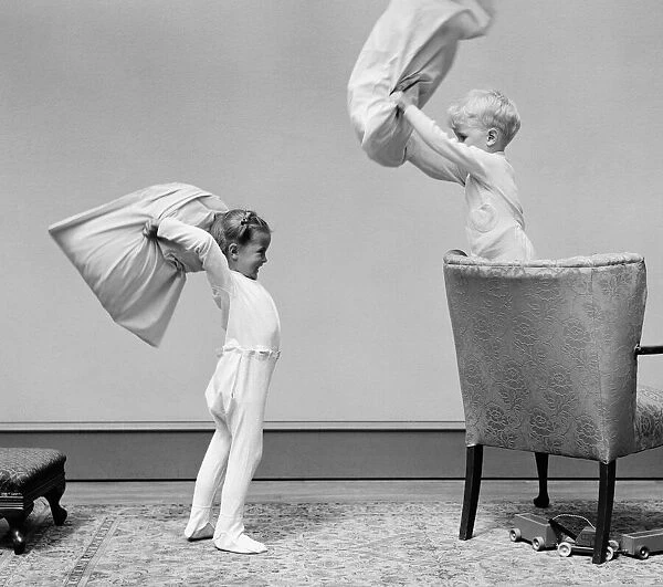 Boy and girl having a pillow fight, boy standing on chair swinging pillow, girl on floor