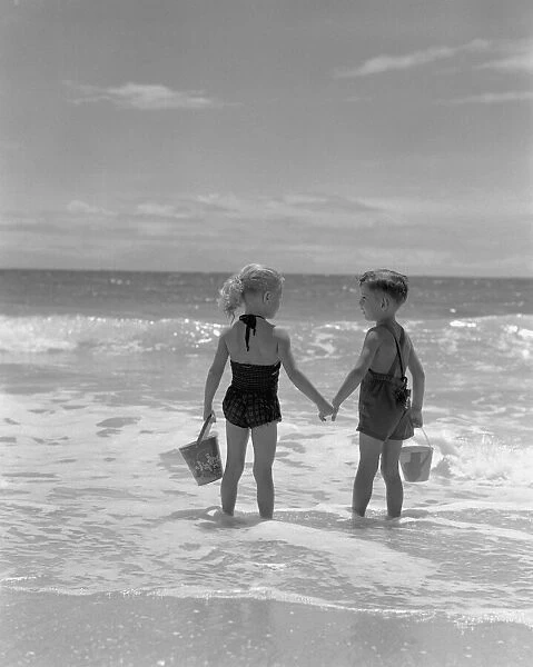 Boy and girl standing on beach, holding hands, rear view