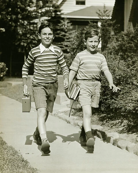 Two boys (10-11) walking side by side in suburbs, carrying books, (B&W)