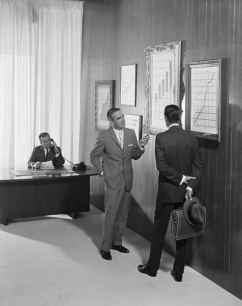 Two businessman discussing at bar chart while another man using telephone in background