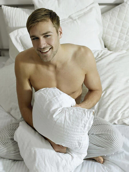 Charming bare chested man wearing pyjamas sitting on a bed, smiling