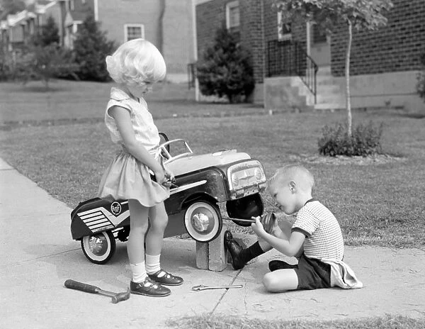 Children on suburban sidewalk, boy playing as mechanic, oiling toy pedal car