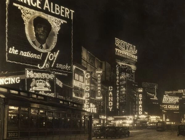 circa 1926: Automobiles and a streetcar pass illuminated theater marquees and billboards