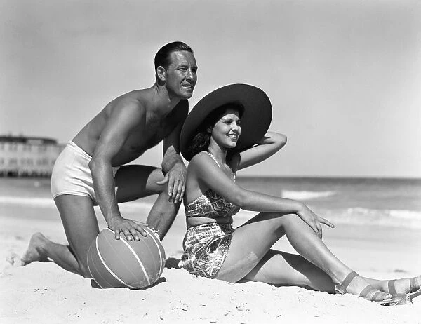 Couple on beach, man leaning on ball, woman sitting look out to ocean, wearing sun hat