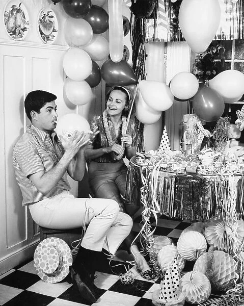Couple blowing up balloons in kitchen for party, (B&W)