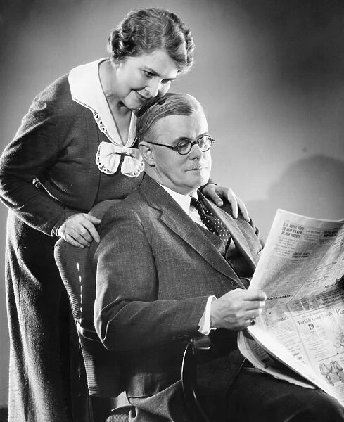 Couple at home, man reading newspaper, woman standing behind, (B&W)