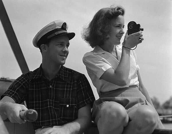 Couple on sailing boat, man holding tiller, woman filming with movie camera. (Photo by H