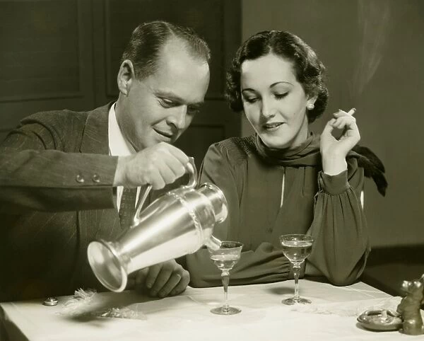 Couple sitting at table, man pouring drinks, (B&W)
