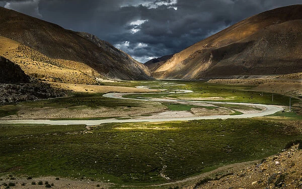 Curves. Meandering creek over grassland and cloudy black sky in Tibet