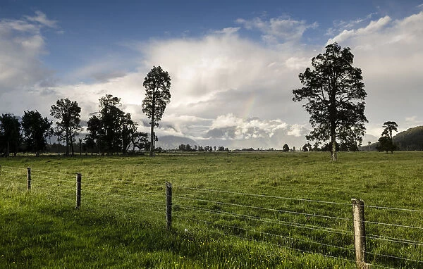 Electric fence and a large sheep pasture, against the backdrop of the Southern Alps, Fox Glacier, storm clouds with a rainbow, South Island, New Zealand