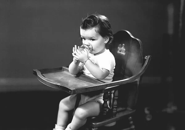 Girl (2-3) sitting in high chair, drinking from glass, (B&W)