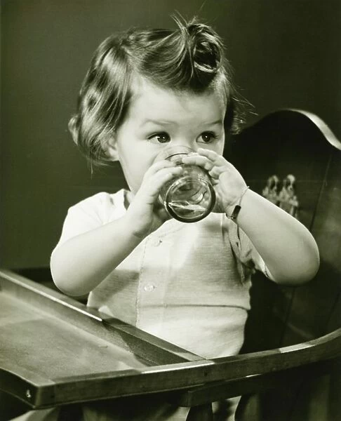 Girl (2-3) sitting in high chair, drinking water from glass, (B&W)