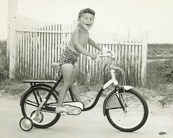 Girl (6-7) riding bicycle with side wheels, (B&W)