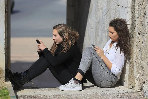 Two girlfriends, teenagers, sitting on the floor, leaning against a wall and writing text message on their mobile phones, Menton, Provence-Alpes-Cote dAzur, France