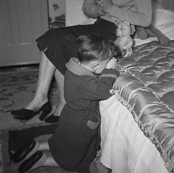 God Bless. 23rd August 1956: A mother sits on a bed to hear her young son in dressing gown