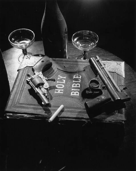 Holy Gun Smoke; Two hand guns and a cigarette lying on a copy of the Holy Bible in a production of Edgar Wallaces play On The Spot