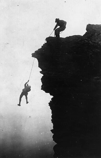 Long Haul. circa 1900: A rock climber swinging on a rope as he is hauled up a cliff face