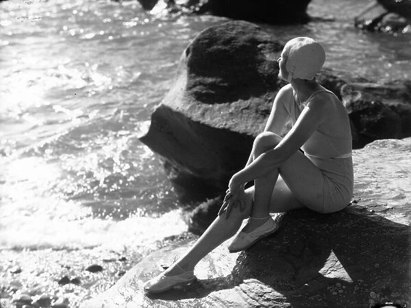 Lorelei. circa 1930: A swimsuited woman sits on a rock gazing out to sea