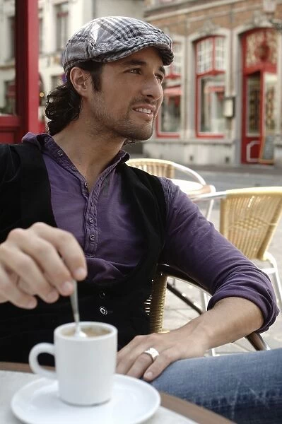 Man, early 30s, wearing casual clothes sitting in an outdoor cafAzA stirring a coffee cup, smiling