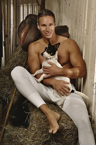 Man with a naked torso wearing long underwear with a dog in a horse barn sitting on bales of hay