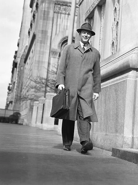 Man in overcoat and hat walking on sidewalk, (B&W), low angle view