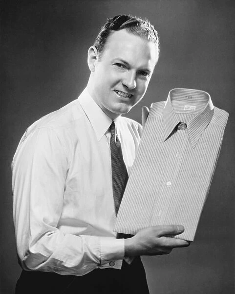 Man presenting folded shirt in studio, (B&W), portrait