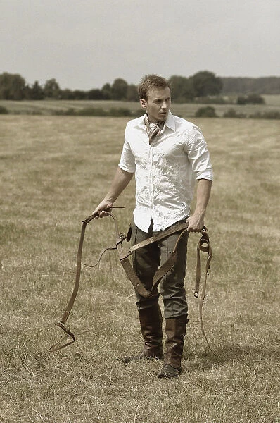 Man wearing riding clothes standing in a field in summer holding a leather strap